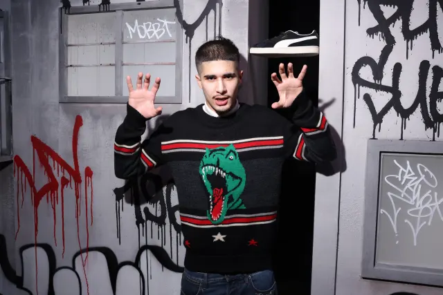 fakemink in a black T-rex graffiti-print sweater, hands raised, in front of a tagged-up set wall